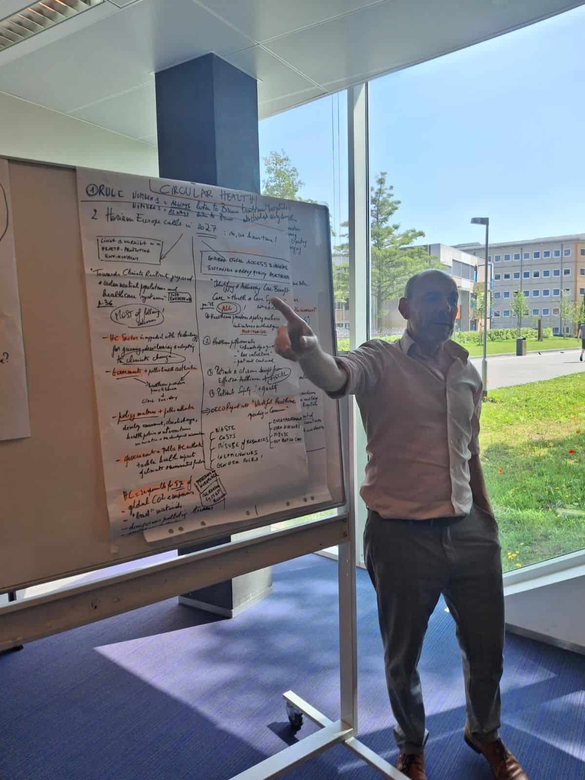 Man pointing at a flipchart covered with handwritten notes in a bright room. Outside, a sunny view with trees and a modern building. Engaged, informative setting.