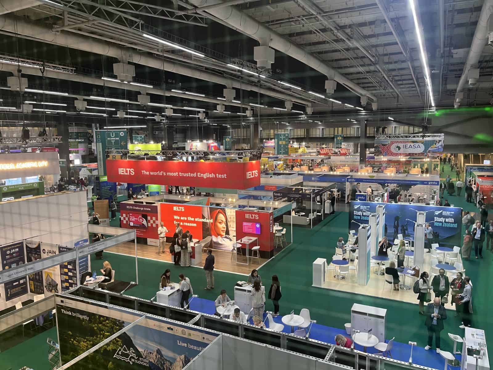 A busy exhibition hall with numerous booths promoting educational services. The prominent red IELTS booth stands out, surrounded by people conversing. The atmosphere is vibrant and professional.