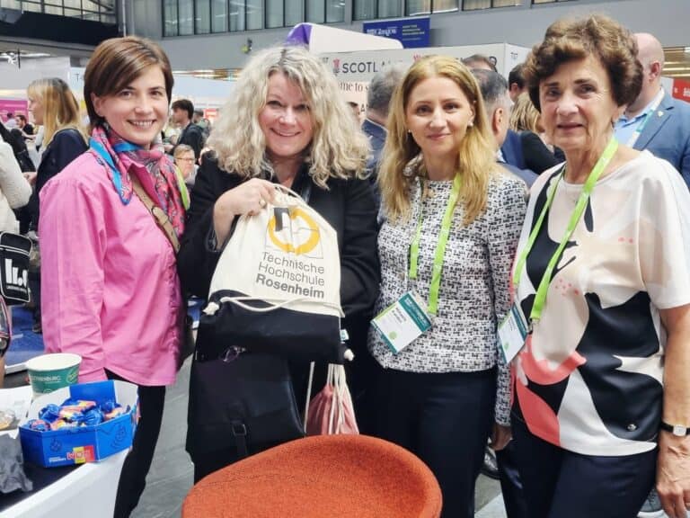 Four women pose at a bustling trade event, smiling warmly. One holds a "Technische Hochschule Rosenheim" bag. The setting is lively and professional.