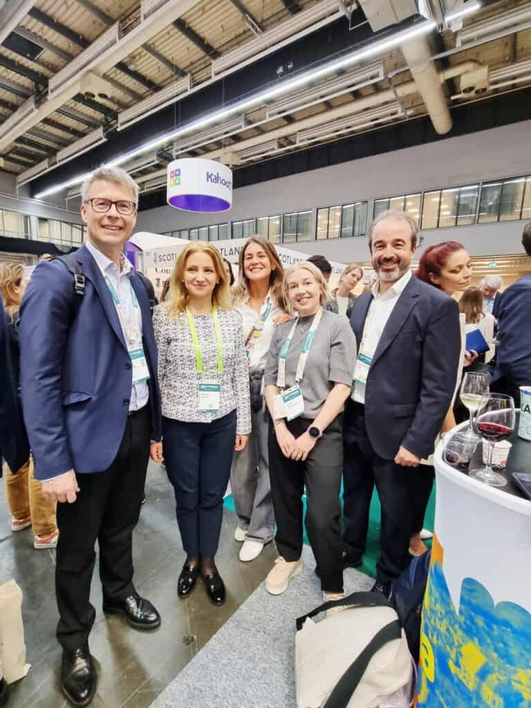 A group of five smiling people stands together indoors at a conference. They wear lanyards, suggesting a professional event. The atmosphere is lively.